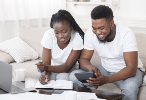 Young happy african american couple planning family budget together, sitting on sofa at home, using calculator and taking notes